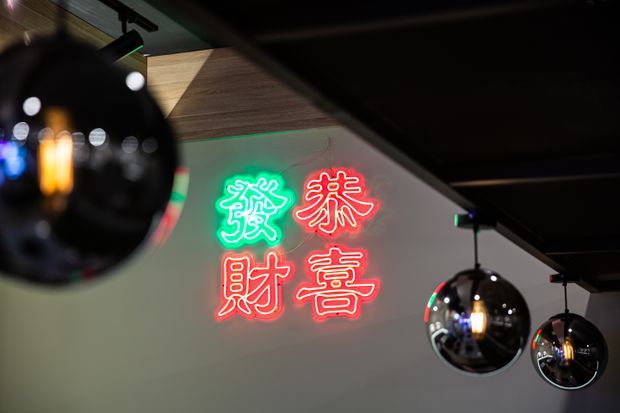 Close-up interior view of a dark, atmospheric commercial space featuring hanging mirrored spherical pendant lights and a colourful Chinese-character neon wall sign in green and red. A timber-lined ceiling section adds warmth against the otherwise dark ceiling and pale wall.