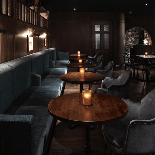 A dark, intimate hospitality lounge or bar interior with round dark timber tables, candle-like table lamps, deep upholstered banquette seating, curved lounge chairs, dark wood wall panelling, a textured dark ceiling, and warm wall sconces. Decorative patterned glazing and an arched opening add visual interest, creating a moody, refined atmosphere.