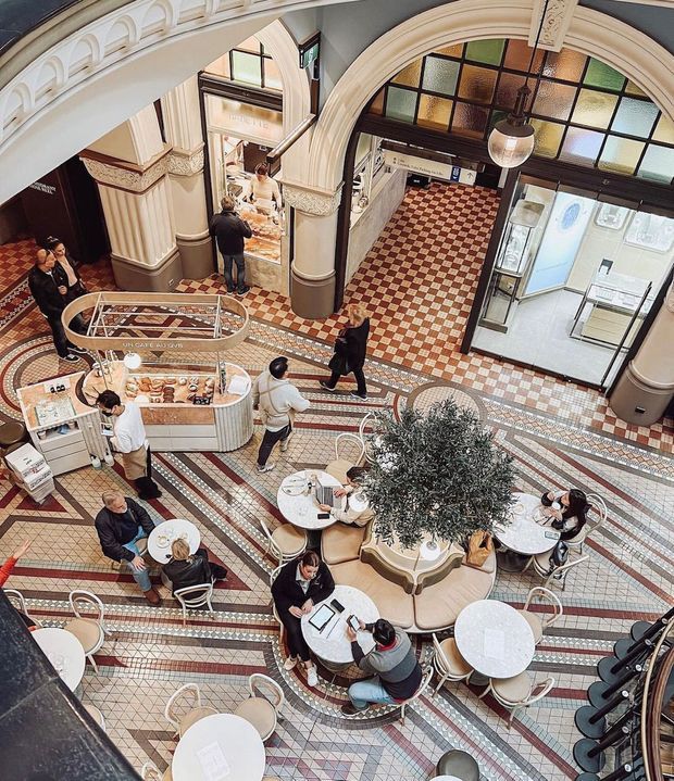 High-angle view of an ornate indoor commercial food hall or café, with patterned mosaic tile flooring, arched openings, classical columns and cornices, stained-glass transoms, café seating, a central circular planter, and food service counters. The space is busy with patrons and staff.