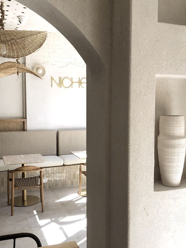 A softly lit, minimalist commercial lounge or reception area viewed through a large rounded archway. The space features textured white plaster walls, a pale upholstered banquette, small stone-topped tables, woven pendant lights, a tiled lower wall, and a sculptural ceramic vase displayed in a recessed niche. A gold wall sign reading “NICHE” is visible in the background.