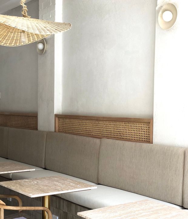A softly lit contemporary dining or lounge area with a long upholstered banquette, pale stone-topped tables, woven cane wall panels, rattan pendant lighting, and rounded wall sconces. The space has a warm neutral palette and refined natural-texture detailing, likely within a hospitality or commercial interior.