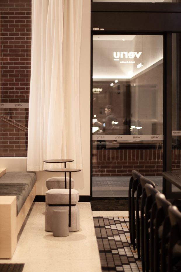 A refined contemporary interior, likely a hospitality or commercial lounge and dining space. The room features a built-in upholstered bench, small round nesting tables, sculptural upholstered stools, a long dining table with dark chairs, warm cream flooring, exposed red brick walls, sheer full-height curtains, and black-framed glazed doors. Reflections reveal additional interior lighting and furnishings beyond the glass.