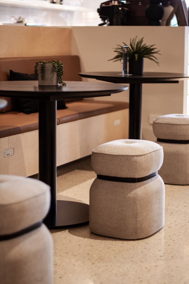 A warm, contemporary café or hospitality interior featuring built-in tan leather banquette seating, round black pedestal tables, upholstered cream stools, light timber joinery, potted plants, and a terrazzo-style floor. The image focuses on the seating and dining area, with a coffee machine visible in the background.