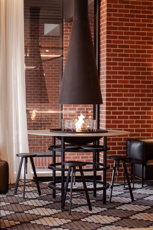 A warm, industrial-style interior centred on a freestanding suspended fireplace over a round high table. The space features exposed red brick walls, full-height glazing with sheer curtains, dark timber and black metal bar stools, a black leather chair, and a geometric patterned floor covering.