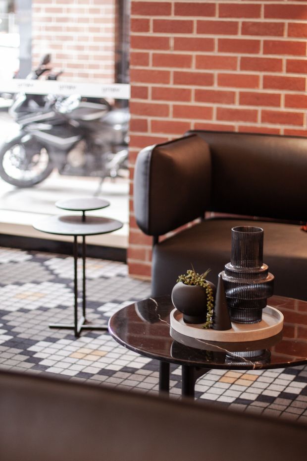 A stylish commercial lounge or reception area with a dark brown leather sofa, round black coffee tables, decorative vessels and a potted plant. A red exposed-brick wall forms the backdrop, while a patterned checkerboard tile floor and large storefront glazing create an urban, industrial atmosphere. Motorcycles are visible outside through the window.