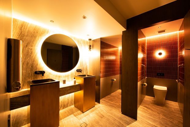 Luxurious, dramatically lit bathroom interior featuring twin pedestal basins, a backlit round mirror, warm wood-toned surfaces, and a separate toilet cubicle clad in dark vertically ribbed tiles. Recessed and concealed LED lighting creates a strong amber glow throughout the space.