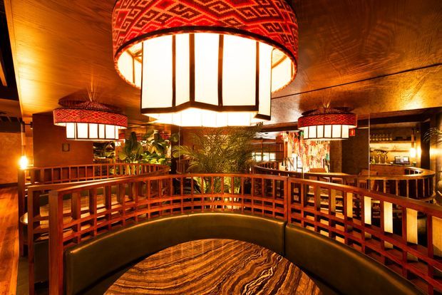 A warmly lit commercial interior with dark timber ceilings, richly stained timber balustrades and panelling, patterned red pendant lamps, and curved built-in banquette seating around a round table. The space has an atmospheric restaurant or lounge character with decorative screens, indoor planting, and a bar or service area visible in the background.