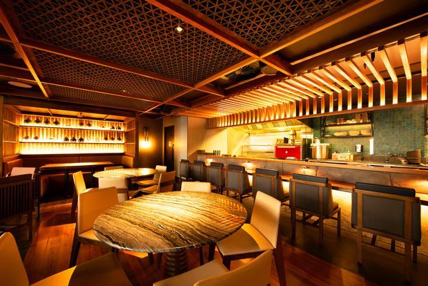 Warm, atmospheric restaurant or bar interior with a long illuminated counter, extensive timber detailing, patterned lattice ceiling, sculptural timber ceiling slats, dark cabinetry, display shelving, and dining tables with upholstered chairs. The space has a refined, intimate hospitality character with strong amber lighting and richly textured wood surfaces.