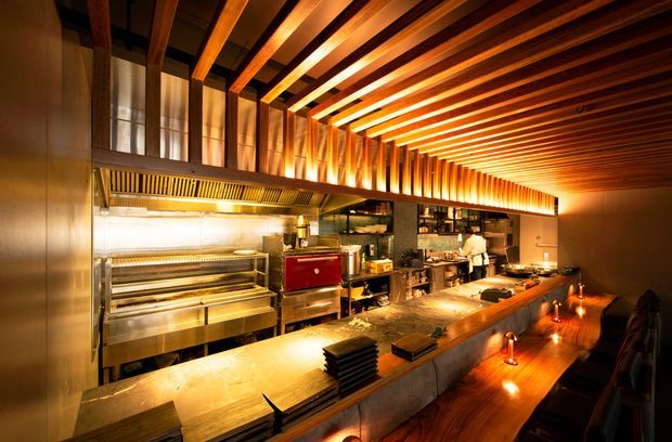 A warmly lit commercial restaurant interior viewed from the customer side of a long bar counter. The open kitchen features stainless-steel cooking equipment, shelving, counters, and staff working behind the bar. Exposed timber ceiling beams and slatted timber detailing create a strong linear architectural feature, while pendant and concealed lighting produce a dramatic amber glow. Dark stools line the timber-fronted counter.