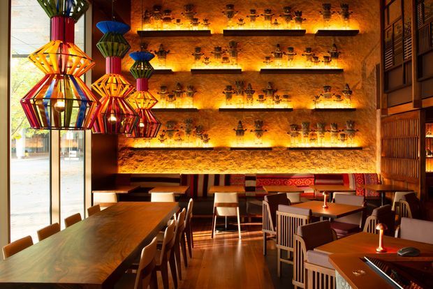 A richly coloured restaurant or bar interior with a warm, atmospheric dining area. The space features dark timber flooring, timber tables and chairs, a textured ochre feature wall with multiple illuminated display shelves holding decorative objects, and bold geometric multicoloured pendant lights. Vertical timber screening and patterned upholstered bench seating add further texture and visual interest.