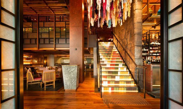 Interior view of a multi-level hospitality venue, likely a restaurant or bar, framed by dark glazed doors. The space features warm timber flooring and ceiling structure, exposed stone and concrete columns, an illuminated multicoloured staircase, dining seating, a bar with bottle display, and a dramatic hanging textile installation.