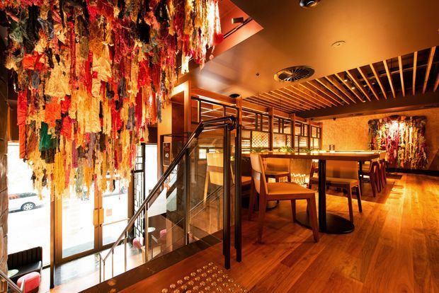Warmly lit upscale restaurant or hospitality interior featuring a bar and dining area, rich timber flooring and ceiling battens, dark timber and glass stair balustrades, patterned timber screens, and dramatic suspended multicoloured textile art forming a prominent feature installation.
