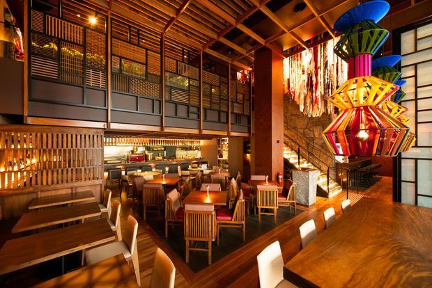 A warmly lit, multi-level restaurant interior with extensive timber ceiling beams and wall slats, dark wood dining tables, upholstered chairs, a visible bar or service counter, and a dramatic cluster of colourful sculptural pendant lights. A mezzanine-style upper level with patterned screens and open railings overlooks the dining area.