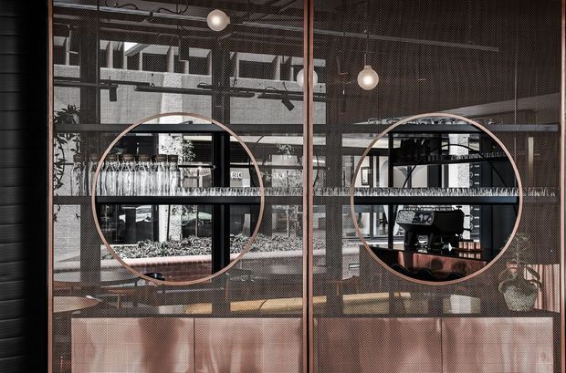 A contemporary commercial café or bar interior viewed through a dark metal mesh or glazed partition. The foreground features copper-toned framed circular openings, glass shelving with rows of wine glasses, and a copper-clad service counter. Behind it are a coffee machine, additional shelving, industrial ceiling services, black-framed glazing, and spherical pendant lights. The composition has a refined industrial character with warm metallic accents and strong geometric detailing.