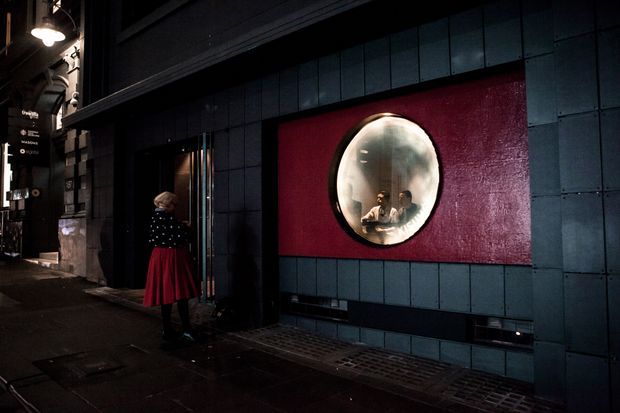 Dark urban streetscape showing the exterior facade of a commercial venue. The building is clad in dark blue-black rectangular panels, with a bold red inset feature panel containing a large circular convex mirror or porthole-like window. A person stands near the recessed entrance, and illuminated signage and industrial-style exterior lighting are visible along the narrow pavement.