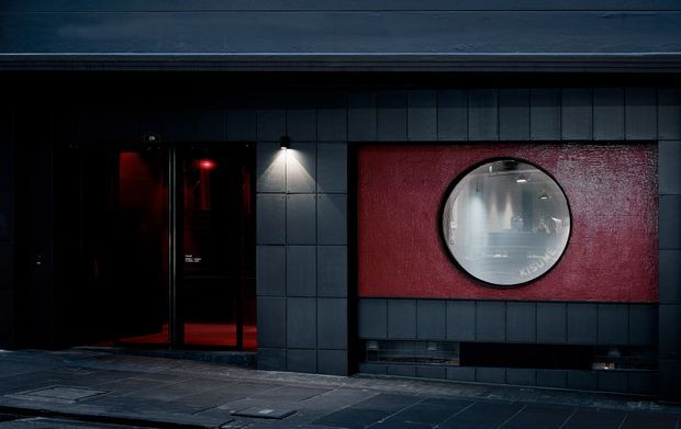 Dark, moody commercial-style building facade clad in dark rectangular panels, with black-framed glazed entry doors, a wall-mounted light, and a red textured feature panel containing a circular window or porthole.