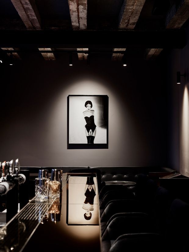 A dark, intimate hospitality-style interior with a glossy black bar counter in the foreground, crystal glassware, and a long black upholstered banquette along the wall. A monochrome framed portrait is dramatically spotlighted against a charcoal wall. The ceiling has exposed rough timber or concrete beams with black-painted services and recessed spotlights, creating a moody, luxurious atmosphere.