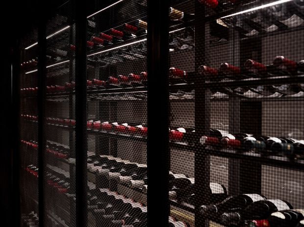 Dark, atmospheric view through black metal mesh wine racks filled with numerous horizontally stored wine bottles. The shelving spans the frame in a compact, high-density storage environment, with black, red, and white bottle capsules providing the main visual accents.