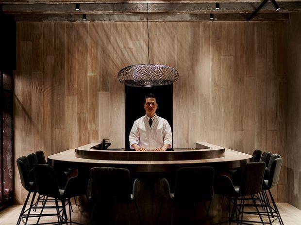 A moody, refined hospitality interior featuring a large curved timber-clad reception or bar counter surrounded by dark upholstered bar chairs. A person stands behind the counter, with a sculptural woven pendant light above. The space has warm wood wall panelling, exposed timber ceiling beams, dark accents, and dramatic directional lighting.
