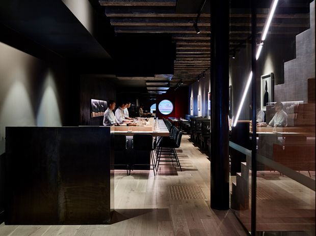 A dark, refined restaurant interior viewed from the entrance or circulation area, with a long central bar and counter seating extending into the space. Diners and staff are visible, along with booth seating, timber ceiling beams, black structural columns, dark wall finishes, tiled flooring, and dramatic linear lighting. Glazing on the right reveals additional seating and reflections within the restaurant.