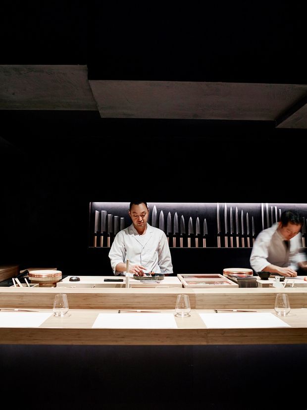 A dark, intimate Japanese-style sushi counter restaurant interior. Two chefs work behind a long pale timber counter with individual white place settings, glasses, plates, and food preparation areas. A backlit display of vertically arranged knife-like forms forms a strong feature wall behind the chefs, while large dark structural ceiling beams and black walls create a dramatic, high-contrast atmosphere.