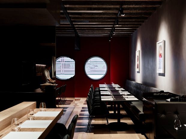 A dramatic, dark-toned restaurant dining interior with rows of neatly set black dining tables and upholstered chairs. The space features a long banquette along a textured dark wall, a bold deep-red partition wall with two circular windows, exposed timber ceiling joists with track lighting, framed artwork, and a pale timber communal table in the foreground. The lighting creates strong pools of illumination and a refined, atmospheric hospitality setting.