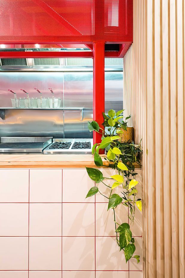 Close-up interior view of a compact commercial-style kitchen or food-service servery. The space features stainless-steel splashback and cooking equipment, a pale tiled cabinet front with contrasting red grout, bright red structural framing and perforated overhead screening, a timber benchtop, and a vertical timber slat wall. Trailing green plants soften the vivid industrial fit-out.