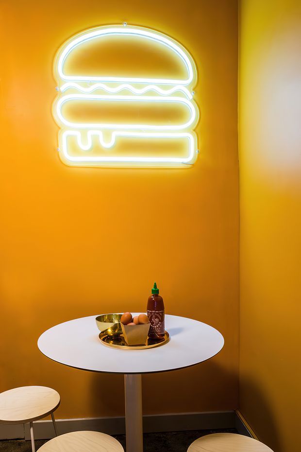 A compact, vividly styled interior corner with saturated orange walls and a glowing white neon hamburger sign. A small round white pedestal table holds a tray, bowl, food items, and a bottle, with pale timber stools around it. Dark flooring and simple skirting frame the warm, playful hospitality setting.