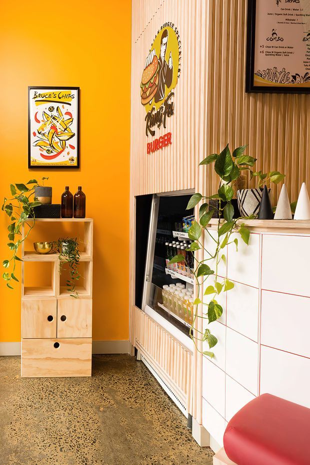 A brightly coloured commercial food venue interior, likely a burger or takeaway shop. The space features a vivid orange painted wall, framed graphic artwork and menu signage, a pale timber vertical slat feature wall with branded burger graphics, an open refrigerated display, terrazzo-style concrete flooring, and a white tiled counter with red grout. Potted trailing plants, dark decorative bottles, and cone-shaped objects add visual interest. The overall design is playful, bold, and retro-inspired.