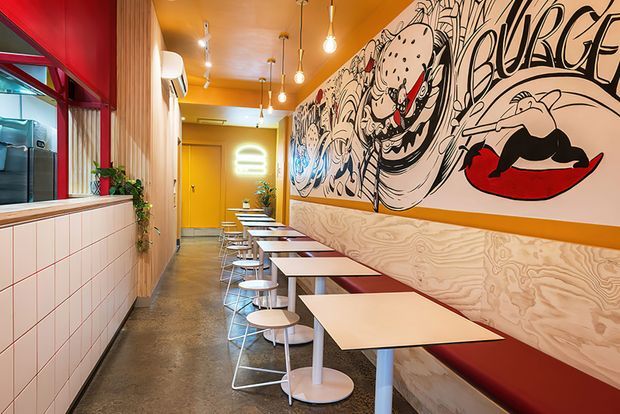 A narrow, brightly coloured commercial dining space, likely a casual restaurant or café. The room features a long banquette with red upholstery, pale wood wall panelling, small square tables with stools, a tiled service counter, warm pendant lighting, and a large black-and-white illustrated mural with red graphic accents. Red-framed glazing and vibrant yellow and red finishes create a bold, playful atmosphere.