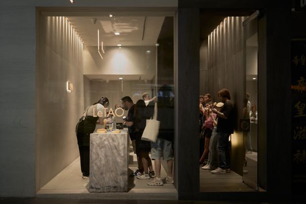 A dimly lit commercial interior viewed through a glazed storefront, showing a narrow contemporary reception or service area. A marble-faced counter stands near the entrance, with several people gathered inside. The space has pale tiled or stone walls, a linear ribbed feature wall with integrated lighting, recessed ceiling lights, and dark framing around the glazing. Chinese lettering is visible on the exterior signage at the right.