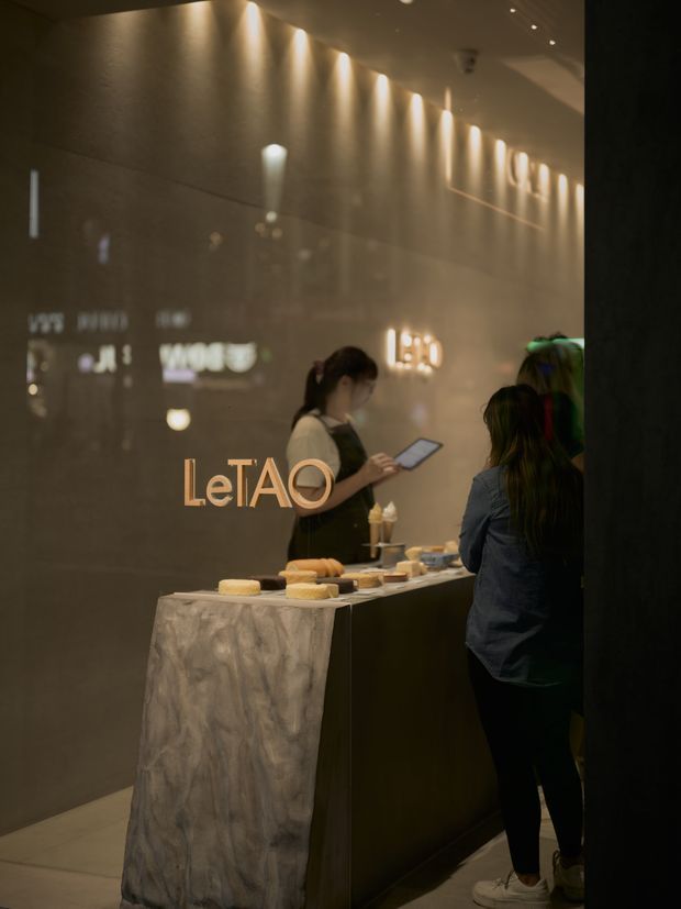 A dimly lit contemporary commercial dessert or food-service counter, with a dark stone or marble-clad island counter, a glossy dark wall, warm linear and spotlight illumination, illuminated signage, and staff serving customers. The image has a refined, intimate hospitality atmosphere.