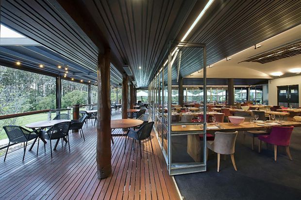 A contemporary restaurant or hospitality dining space with a covered timber deck running alongside floor-to-ceiling glazing. The interior contains numerous dining tables and chairs, while the deck provides additional outdoor seating overlooking a wooded landscape. Dark linear timber ceiling boards, exposed timber posts, glass partitions, warm wood decking, and a dark carpeted dining area create a refined indoor-outdoor setting.