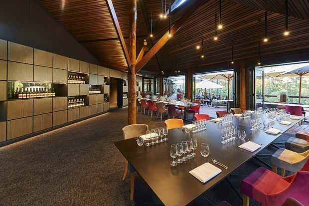 A sophisticated restaurant or hospitality dining interior with a large communal dark dining table in the foreground, set with numerous wine glasses, water glasses, cutlery and folded napkins. The room features a dramatic exposed timber post-and-beam structure, richly patterned timber slatted ceiling, dark walls, integrated bottle display and bar cabinetry, and extensive floor-to-ceiling glazing opening to an outdoor terrace. Colourful upholstered dining chairs add accents of orange, red, purple and blue against the dark, warm-toned interior.