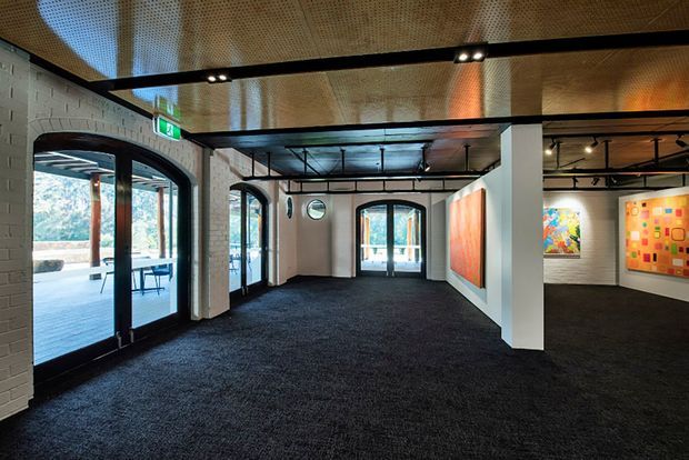 A contemporary gallery or commercial interior with an open, carpeted exhibition space. The room features white-painted brick walls, black steel-framed arched glazed doors and windows, exposed black ceiling beams with track lighting, and a perforated timber-look acoustic ceiling. Several colourful artworks are mounted on the walls, and a glazed entrance opens to an exterior terrace or walkway.
