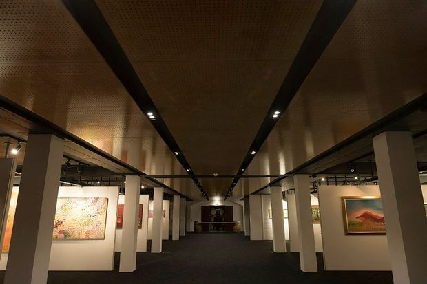 A dimly lit contemporary art gallery interior with a long central passage, white partition walls displaying framed artworks, dark carpet, white structural columns, and a warm perforated timber-toned ceiling with recessed linear lighting.