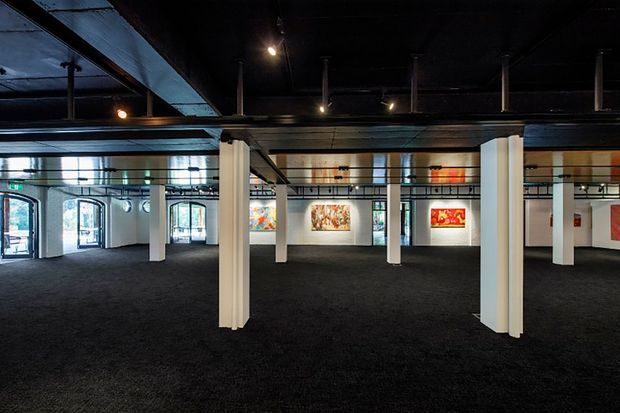 A spacious two-level gallery or exhibition interior with dark industrial ceilings, exposed structural beams and columns, white support pillars, polished concrete or dark carpeted flooring, and artworks displayed on white walls. Large arched windows and glazed doors line the perimeter, while an upper mezzanine with a metal balustrade overlooks the main exhibition floor. The lighting is subdued and atmospheric.