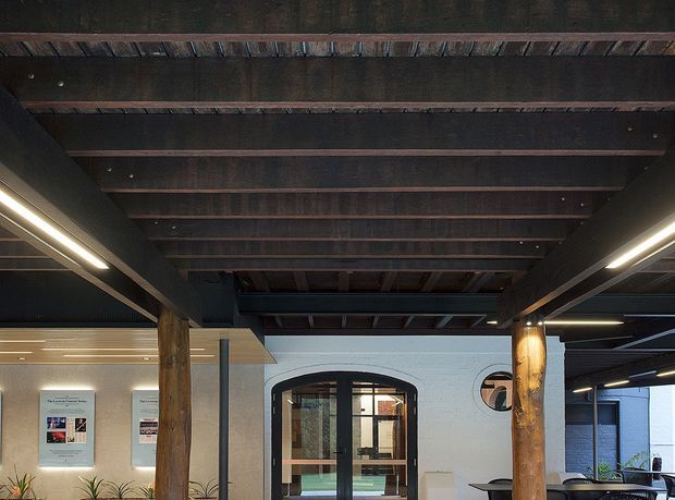 Interior architectural view of a dark timber-beamed ceiling supported by substantial timber posts. Linear strip lighting runs along both sides, illuminating a white painted masonry wall with an arched glazed doorway and circular window. A small reception or seating area, wall-mounted information displays, and indoor planting are visible. The composition highlights exposed structural timber and an industrial, adaptive-reuse character.