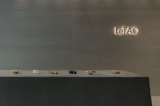 A minimalist commercial display or gallery interior featuring a long dark monolithic counter with small objects arranged on top. Large grey wall panels form the backdrop, with softly illuminated “LeTAO” signage adding a subtle focal point. The space has restrained detailing, low warm lighting, and a quiet, refined presentation.