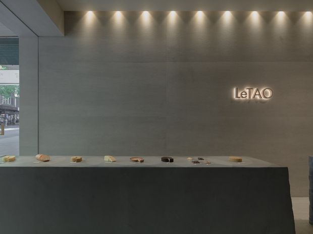 A minimalist commercial retail or hospitality interior featuring a long dark stone counter with small displayed items, a large grey stone or concrete-look feature wall, and a backlit “LeTAO” sign. Recessed spotlights create a refined, subdued atmosphere, while a glazed opening at left reveals the street outside.