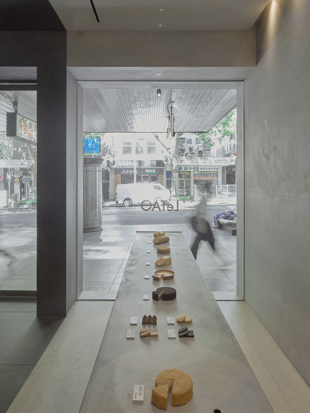 A minimalist retail or gallery-like commercial interior viewed toward a full-height glazed street frontage. The space has raw grey concrete walls and floor, a deep rectangular entry opening, a linear recessed ceiling light, and a long central display plinth presenting small objects or samples. The street and shopfronts are visible through the glass, with a blurred passerby outside. A large mirror or reflective glazing on the left repeats the interior and storefront view.