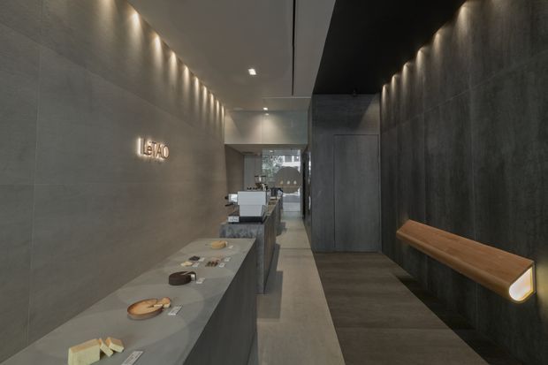 A dimly lit contemporary commercial interior, likely a reception or showroom entry, with a long grey stone counter displaying small product samples, illuminated wall signage, dark textured wall finishes, integrated linear lighting, and a timber wall-mounted bench or handrail. A narrow corridor and glazed opening extend toward the rear.