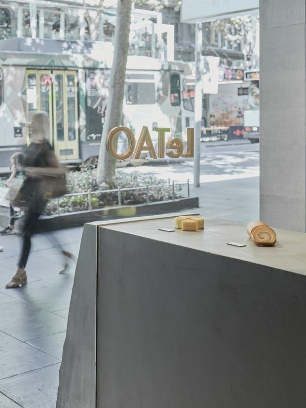 A bright contemporary commercial storefront or gallery interior viewed from inside through a large glazed frontage. A substantial dark grey sculptural display plinth or counter occupies the foreground, holding two small sculptural or product objects. Reverse lettering is mounted on the glazing, with a busy urban pedestrian streetscape, tram, trees, and neighbouring shopfronts visible outside. The space has a minimalist, gallery-like presentation with concrete-look surfaces and abundant daylight.