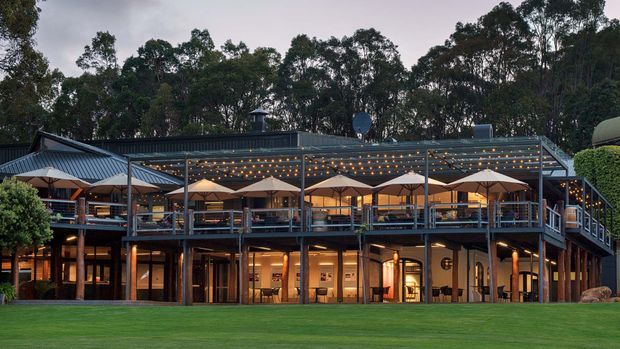 Exterior view of a large two-storey hospitality or commercial building set against dense woodland. The building has a broad low-pitched metal roof, extensive glazed openings, dark timber or steel structural posts, and a prominent elevated upper terrace lined with outdoor dining tables, umbrellas, railings, and festoon lighting. Warm interior lighting glows through the lower-level windows, with a manicured lawn in the foreground.