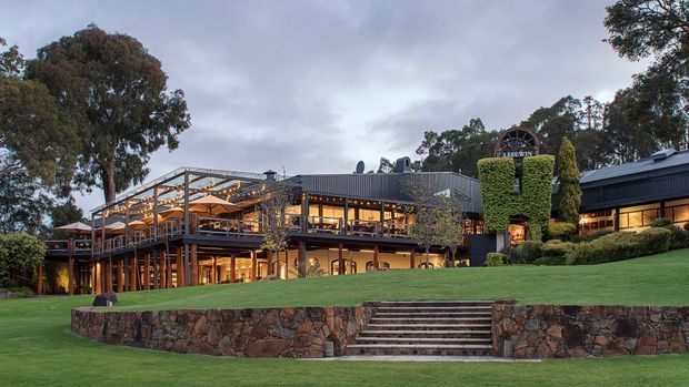 A large multi-level commercial or hospitality building set within landscaped grounds, photographed at dusk. The structure features extensive warm-lit glazing, dark metal cladding, exposed timber or steel framing, broad outdoor terraces, and a prominent stone retaining wall with central stairs leading across the lawn.