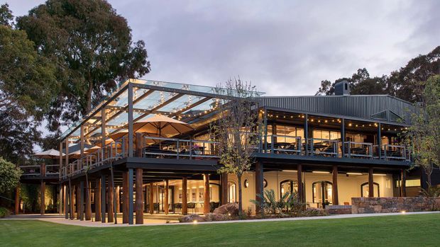 A large two-storey contemporary building set among mature trees, photographed at dusk. The structure features dark metal cladding, extensive glazing, a broad timber-and-steel pergola over an elevated outdoor terrace, warm integrated lighting, and stone masonry at the lower level. The illuminated interiors and terrace suggest a hospitality or commercial venue, with landscaped lawn in the foreground.