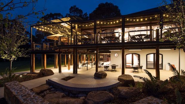 Night-time exterior view of a multi-level residence or hospitality-style building with expansive covered terraces, dark timber or metal posts, warm string lighting, outdoor dining and lounge furniture, and illuminated arched openings on the lower level. The building is integrated into a landscaped rocky garden with paved paths and raised deck areas.