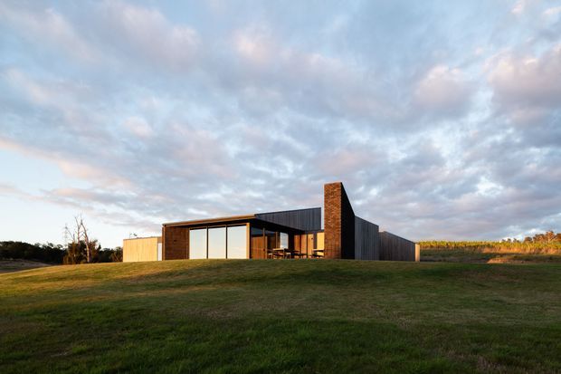 A low-profile contemporary single-storey residence set in an open rural landscape. The building has dark vertical cladding, a prominent exposed brick chimney, extensive glazed openings, and a simple flat or very low-pitched roof form. A broad lawn and agricultural fields surround the house beneath a dramatic cloudy sky.