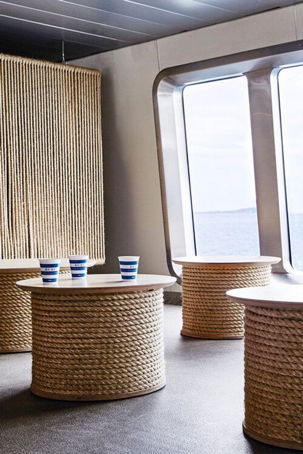 A bright ferry or boat interior with large rounded rectangular windows overlooking the water. Cylindrical tables and seating are wrapped in thick natural rope, paired with pale round tabletops and a matching rope-textured wall panel. The space has a compact marine-inspired fit-out with grey flooring, light wall panels, and metallic window surrounds.