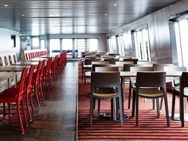 Bright contemporary dining area, likely inside a ferry or other transport vessel, with rows of tables and dark chairs, a long red bar-height seating arrangement on the left, large windows along the sides, and a ribbed metallic ceiling and wall finish. The space has a practical commercial/public interior layout with strong red and neutral contrasts.